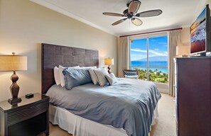 A hotel room with a large bed, a ceiling fan, a nightstand with a lamp, a TV, and a view of the ocean.