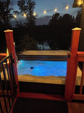 Un jacuzzi con terraza de madera y barandas, rodeado de árboles y luces colgantes.