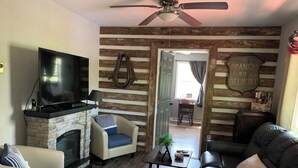 A living room with a fireplace, a television, and a ceiling fan. There is a door leading to another room.