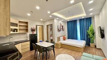 A modern hotel room with a bed, a dining table, a kitchen area, and a large window with curtains.