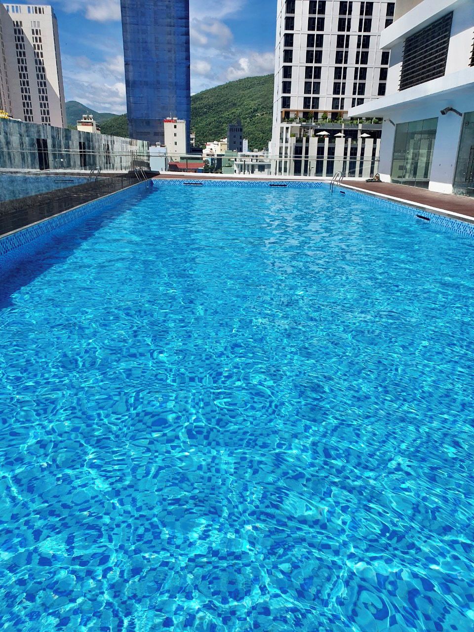 A swimming pool with clear blue water, surrounded by modern buildings and a mountainous backdrop.