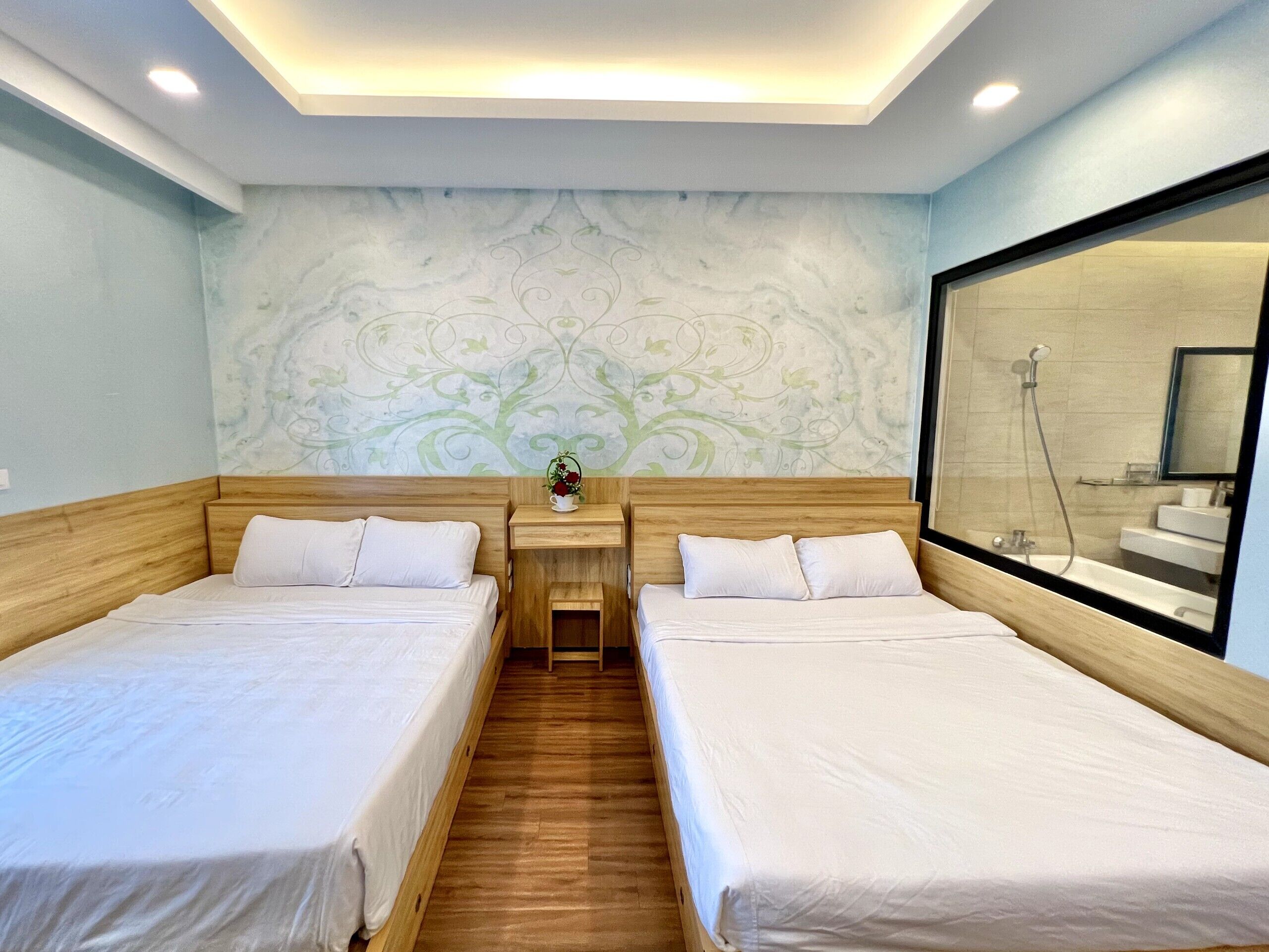 Habitación de hotel con dos camas individuales, una mesita de noche con un florero y un espejo grande. Se ve una zona de ducha a través de la puerta de cristal.