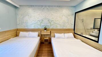 Habitación de hotel con dos camas individuales, una mesita de noche con un florero y un espejo grande. Se ve una zona de ducha a través de la puerta de cristal.