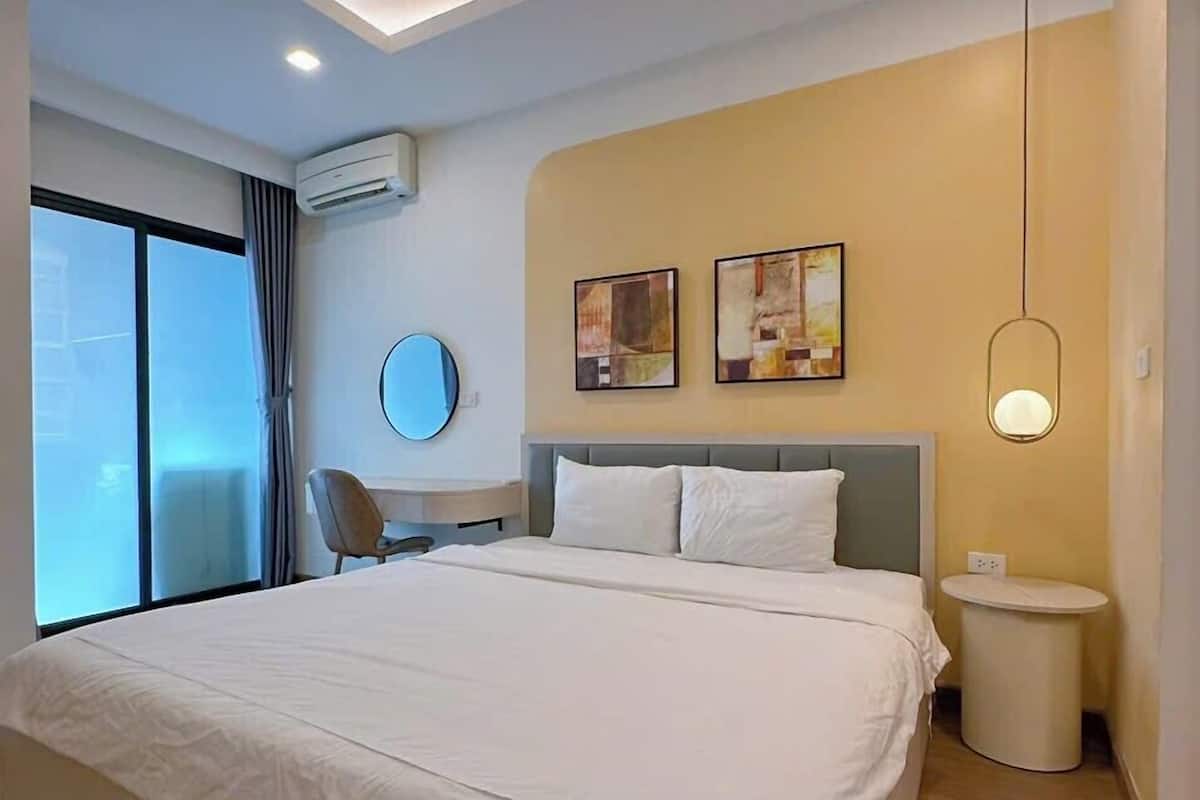 A modern hotel room with a large bed, a round mirror, two paintings, a bedside table, a round nightstand, and a window with curtains.