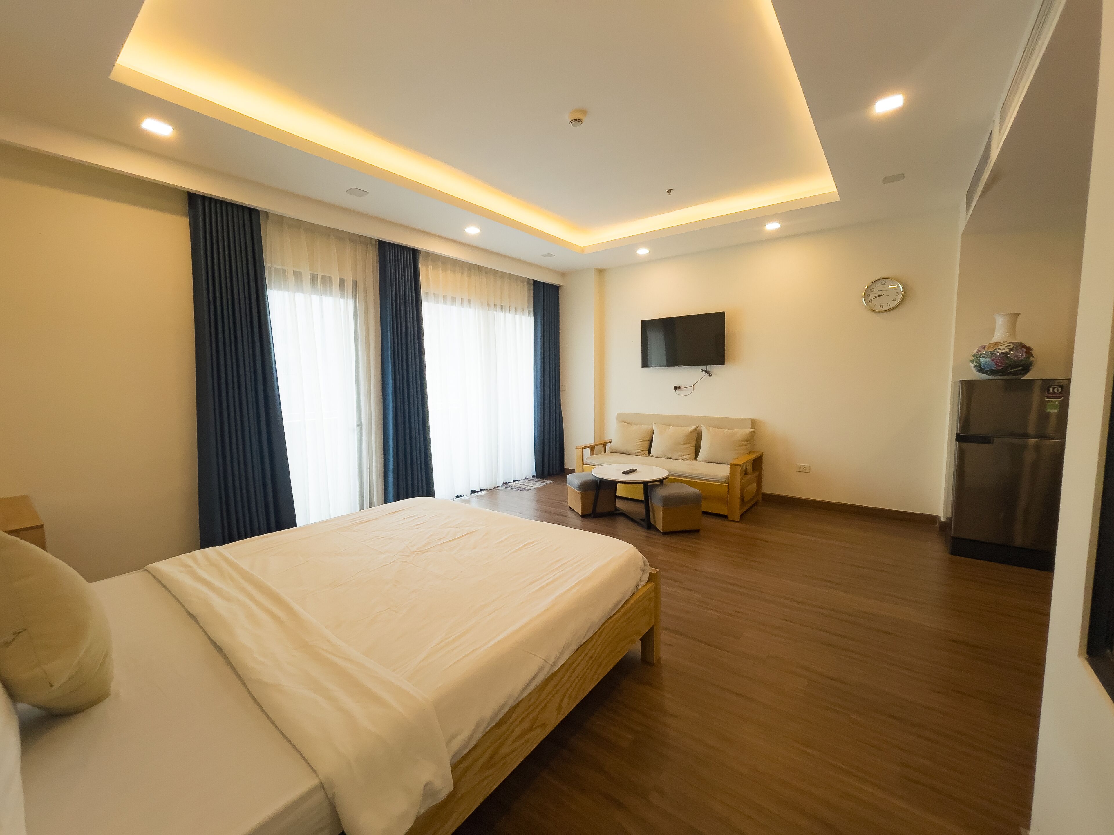 Una habitación de hotel moderna con una cama grande, un sofá, una mesita y un refrigerador. Un televisor está montado en la pared.