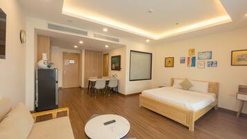 A modern hotel room with a bed, a sofa, a small round table, a dining area with chairs, and a refrigerator.