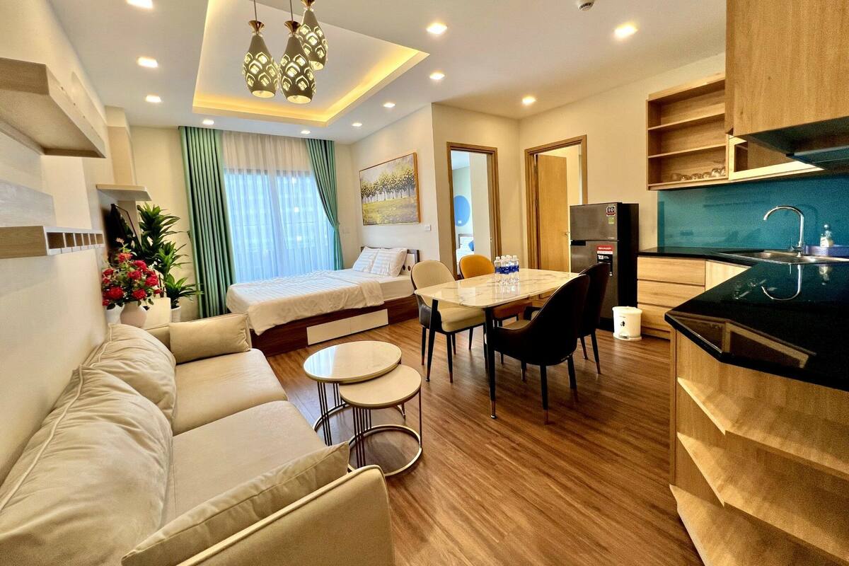 A modern hotel room with a bed, sofa, dining table, chairs, and a kitchen area.