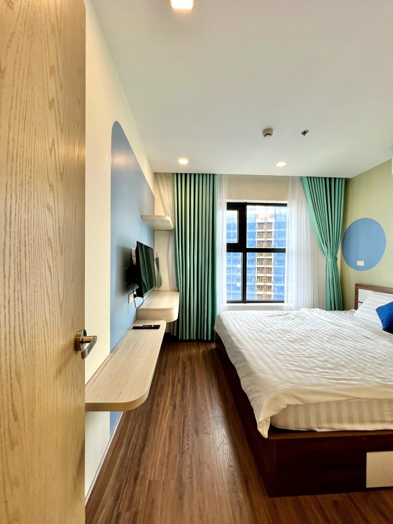 Una habitación de hotel moderna con suelo de madera, una cama con ropa de cama blanca, un televisor montado y una ventana con cortinas.