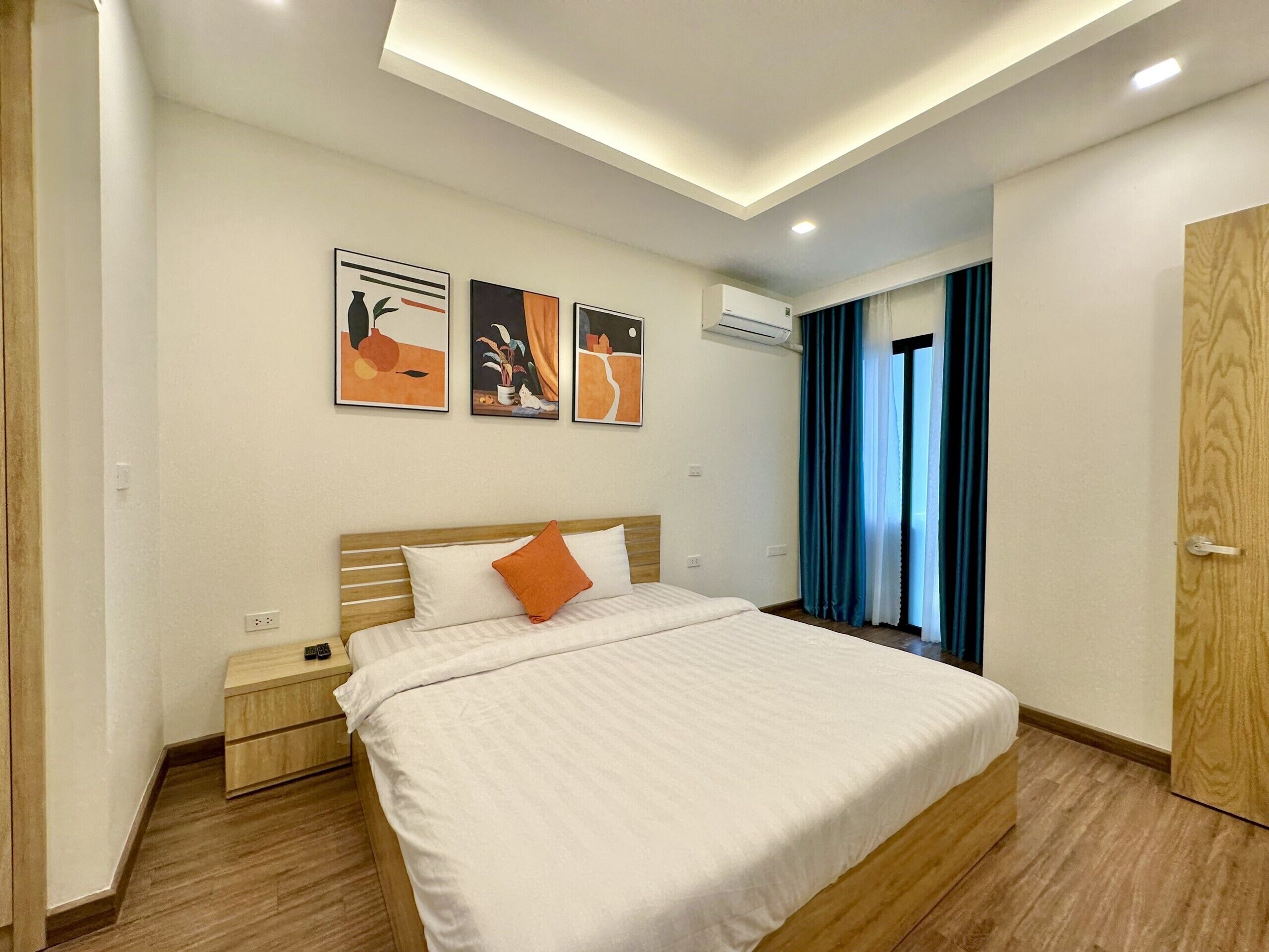 A modern hotel room with a large bed, a nightstand, and a wooden wardrobe. There are three wall-mounted artworks and an air conditioning unit above the bed.