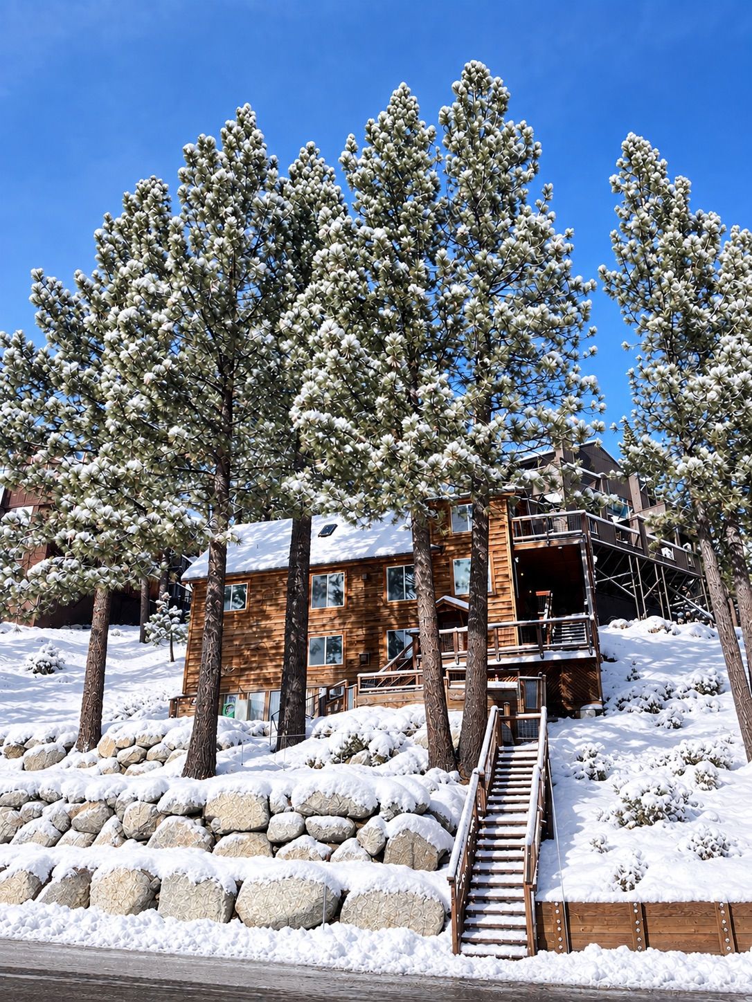 A wooden cabin with a staircase leading up to a deck, surrounded by snow-covered trees and rocks.