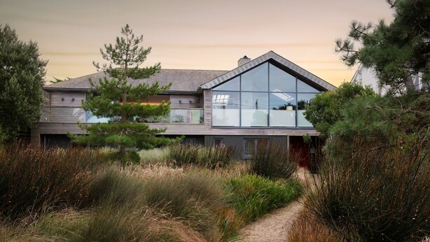Una casa moderna con un amplio ventanal, rodeada de vegetación natural y un camino de grava.