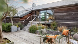 Una casa moderna con fachada de madera, una escalera que conduce a una puerta de cristal y mobiliario exterior con sillas y cojines.