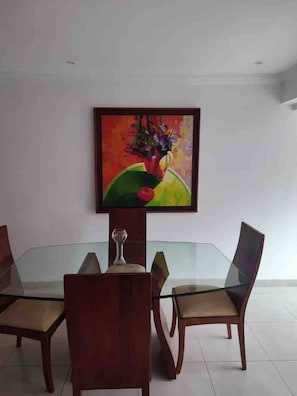 Uma área de refeições com uma mesa de vidro rodeada por cadeiras de madeira, um quadro acima da mesa e uma taça de vinho sobre ela.