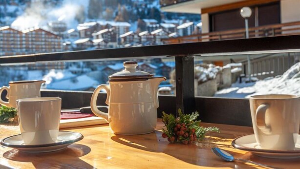 Una mesa con un juego de té, dos tazas y una pequeña planta, con vistas a un pueblo montañoso nevado.