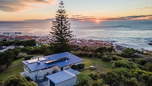 Une maison moderne avec un toit bleu, entourée d’une végétation luxuriante et bordée par une côte rocheuse.