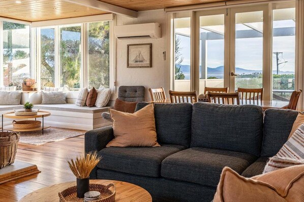 A spacious living room with a dark grey sofa, a wooden coffee table, and a large window offering a view of the outdoors. - Beach Path House - Bicheno Accommodation (Bicheno)