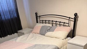 A bed with a metal frame, a bedside table with drawers, and a window with a black curtain.