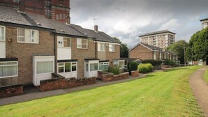 A row of terraced houses with front gardens, a paved walkway, and a large building in the background.