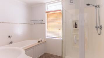 Een badkamer met een bad, een douche met glazen wanden en een raam met zonwering.