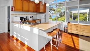 A kitchen with wooden cabinets, a large island with stools, a view of the sea, and a white ceiling. - On the Rocks Bicheno - Bicheno Accommodation (Bicheno)