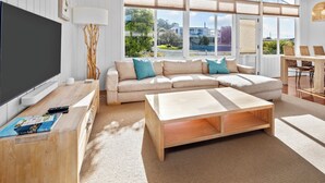 A modern living room with a large TV, a sofa, a coffee table, and a dining area with wicker chairs. - On the Rocks Bicheno - Bicheno Accommodation (Bicheno)