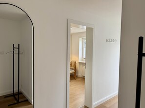 A hallway with a coat rack, a door leading to a room with a chair and a radiator, and a window with blinds.