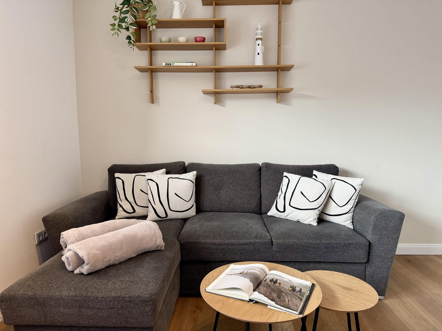 Een moderne woonkamer met een grijze modulaire bank, twee bijpassende sierkussens, een klein bijzettafeltje met een tijdschrift en een houten wandplank boven de bank.