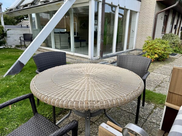 A round wicker table with four chairs, a glass sliding door, and a brick house in the background.