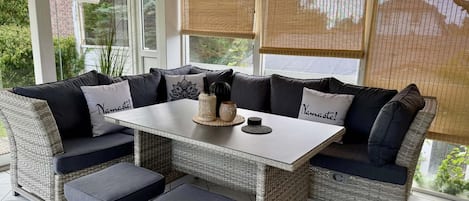 A sunlit outdoor seating area with wicker furniture, a white table, and a view of greenery.