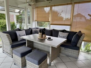 A sunlit outdoor seating area with wicker furniture, a white table, and a view of greenery.