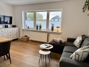 A modern living room with a sofa, a small table, a TV, and a window with plants.