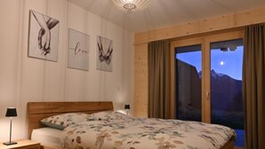 Un dormitorio con una cama de madera, una mesita de noche, una lámpara y vistas a las montañas a través de una puerta corredera.
