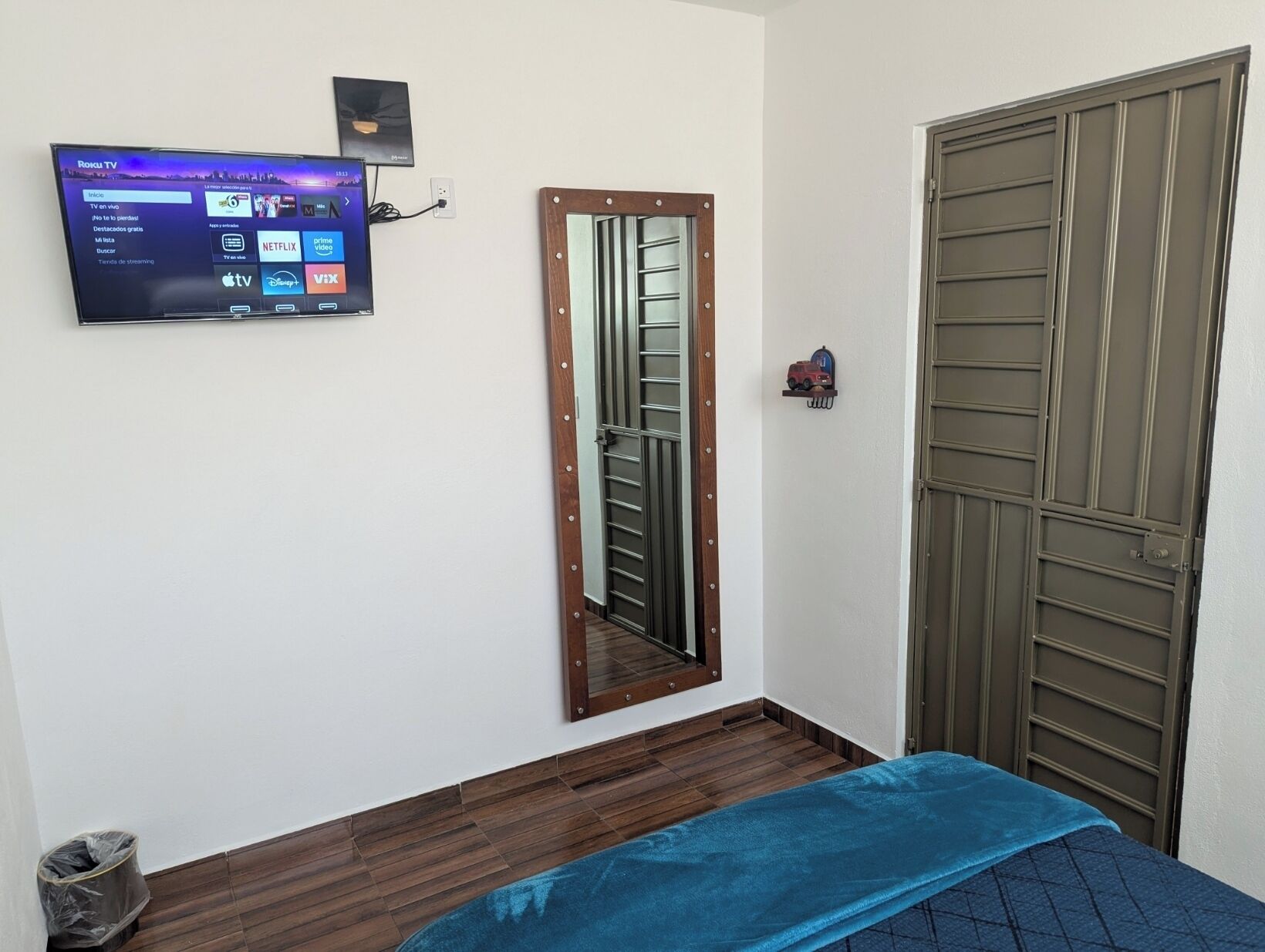 Una stanza con una TV a muro che mostra l'interfaccia di un servizio di streaming, una porta in legno con una griglia metallica e una coperta blu.