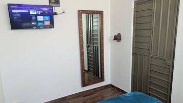 Una stanza con una TV a muro che mostra l'interfaccia di un servizio di streaming, una porta in legno con una griglia metallica e una coperta blu.
