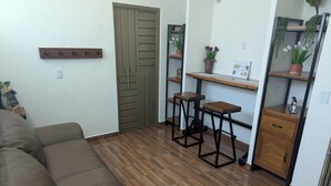 A living room with a brown sofa, a wooden floor, a green door, and a small kitchenette with a wooden table and stools. - Moderna, Comoda Suite Palma en Tepotzotlán centro (Tepotzotlán)