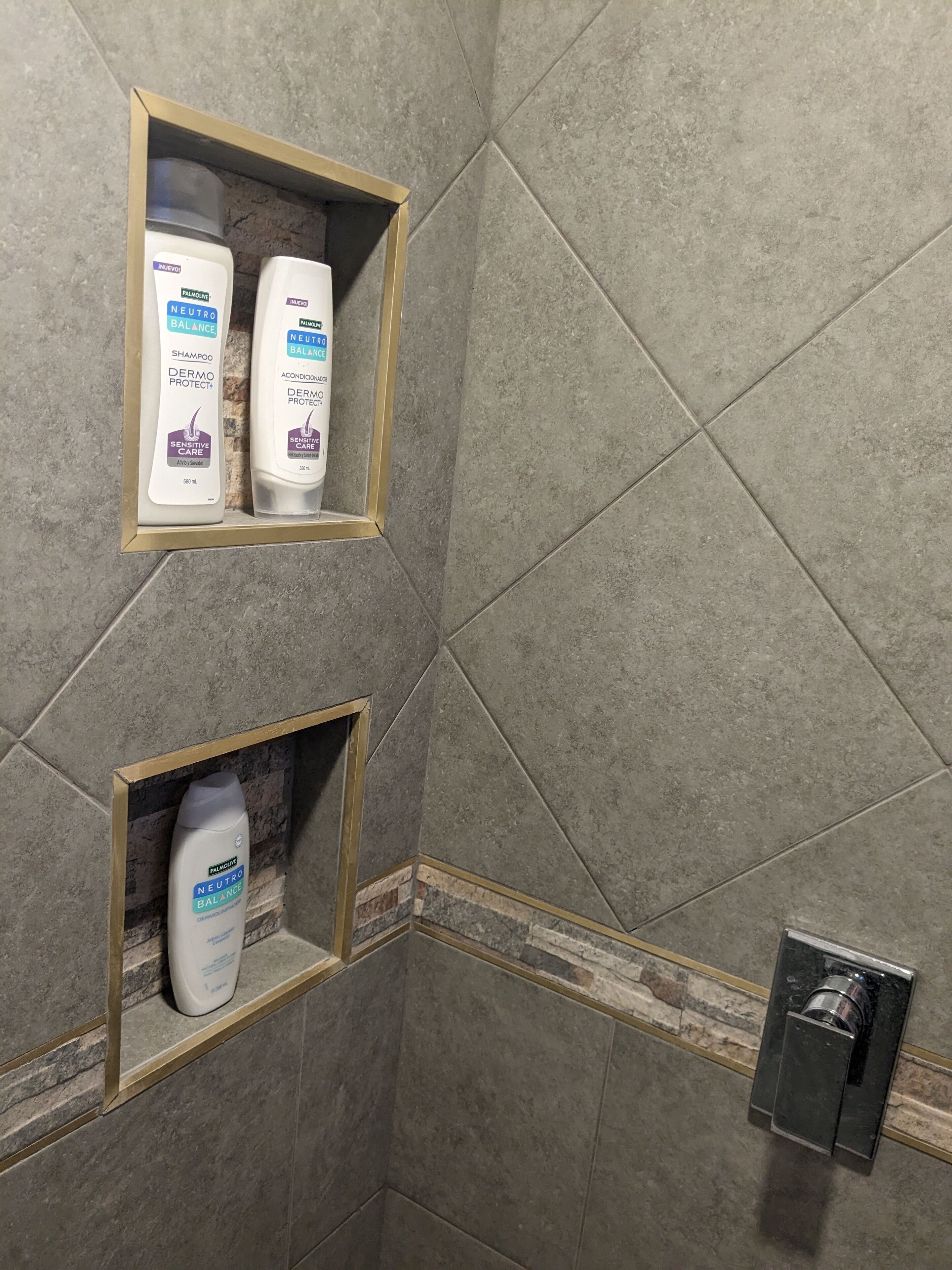 Ein Badezimmer mit grauen Fliesenwänden, zwei Ablagen mit Pflegeprodukten und einem Duschkopf.