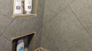 Ein Badezimmer mit grauen Fliesenwänden, zwei Ablagen mit Pflegeprodukten und einem Duschkopf.