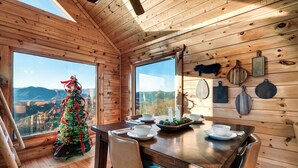 A wooden cabin interior with a dining table, chairs, a Christmas tree, and a large window overlooking a mountainous landscape. - Amazing Mtn views/Hot tub/Fireplace/Bunk beds/Arcade games (Sevierville)