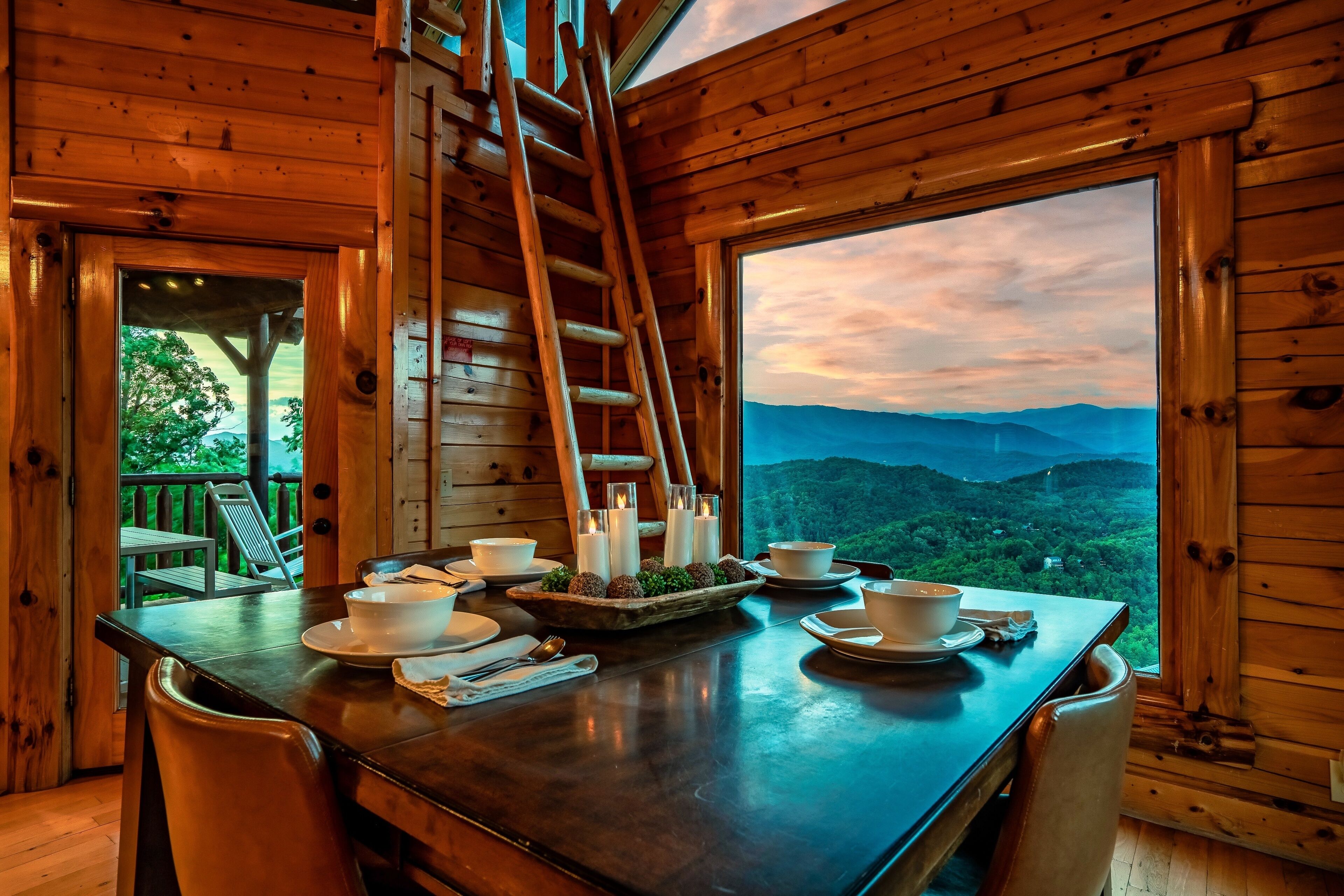 A wooden cabin with a dining table set for two, a ladder, and a scenic mountain view.