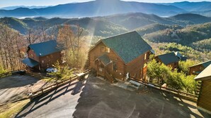 A log cabin with a green roof, surrounded by a parking area and a wooden fence, overlooking a mountainous landscape. - Amazing Mtn views/Hot tub/Fireplace/Bunk beds/Arcade games (Sevierville)
