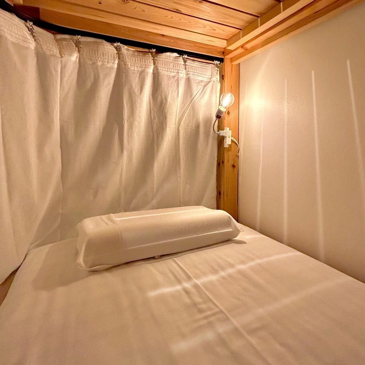 Un lit simple avec un matelas et un oreiller blancs, dans une pièce dont le plafond est orné de poutres en bois et les murs sont peints en blanc.