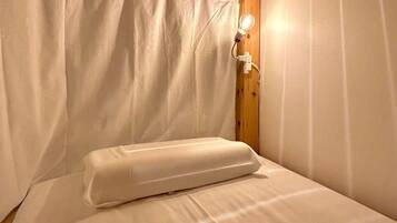 Un lit simple avec un matelas et un oreiller blancs, dans une pièce dont le plafond est orné de poutres en bois et les murs sont peints en blanc.