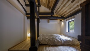 A small, cozy room with a wooden floor, a bed with a white comforter, a window with a view of greenery, and a wall-mounted air conditioning unit. - KOTOBUKIAN (Awaji)