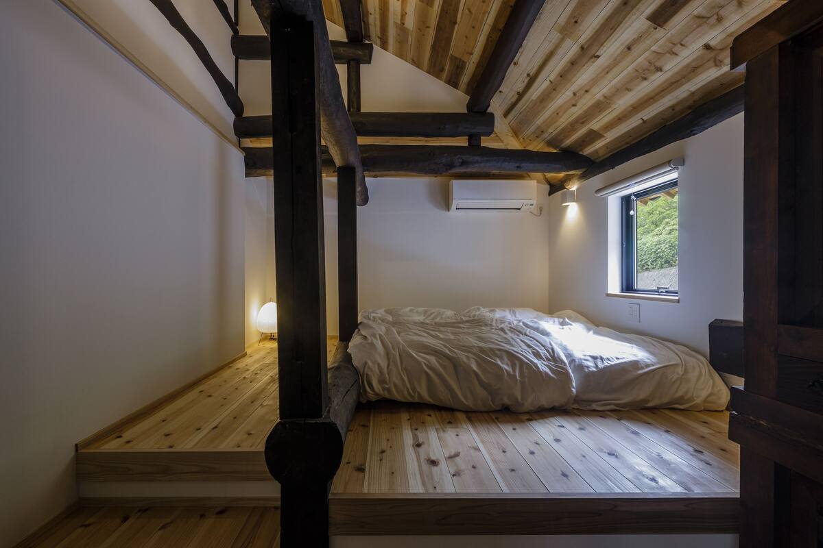 A small, cozy room with a wooden floor, a bed with a white comforter, a window with a view of greenery, and a wall-mounted air conditioning unit.
