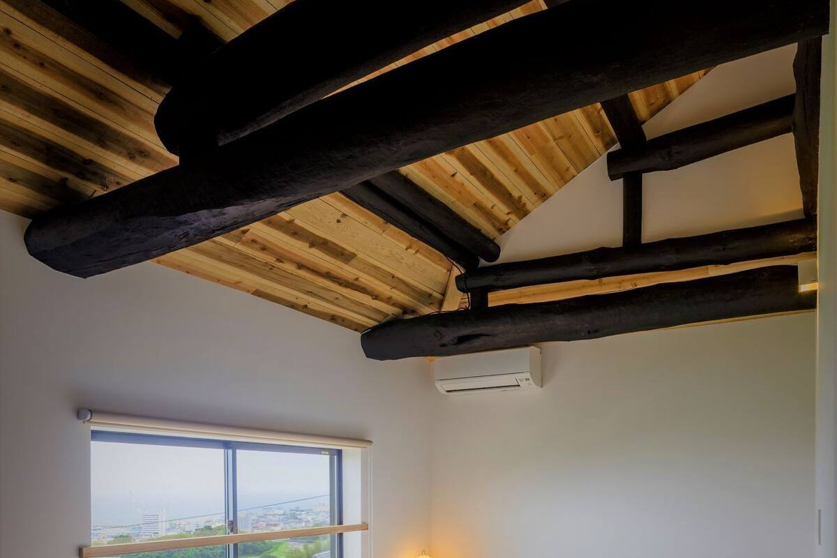 A bedroom with a wooden ceiling, a large window with a view, and a bed with white bedding.