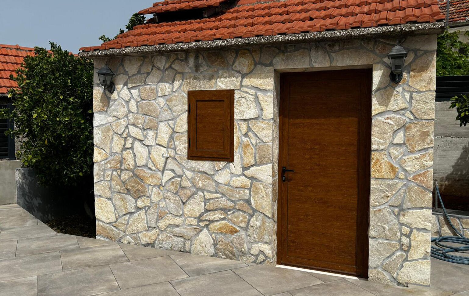 Un edificio de piedra con dos puertas de madera y una ventana pequeña, rodeado de un área pavimentada y follaje.