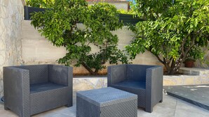 Un espace patio aménagé avec deux canapés en osier gris et un pouf assorti, le tout entouré de verdure et bordé d’un mur en pierre.
