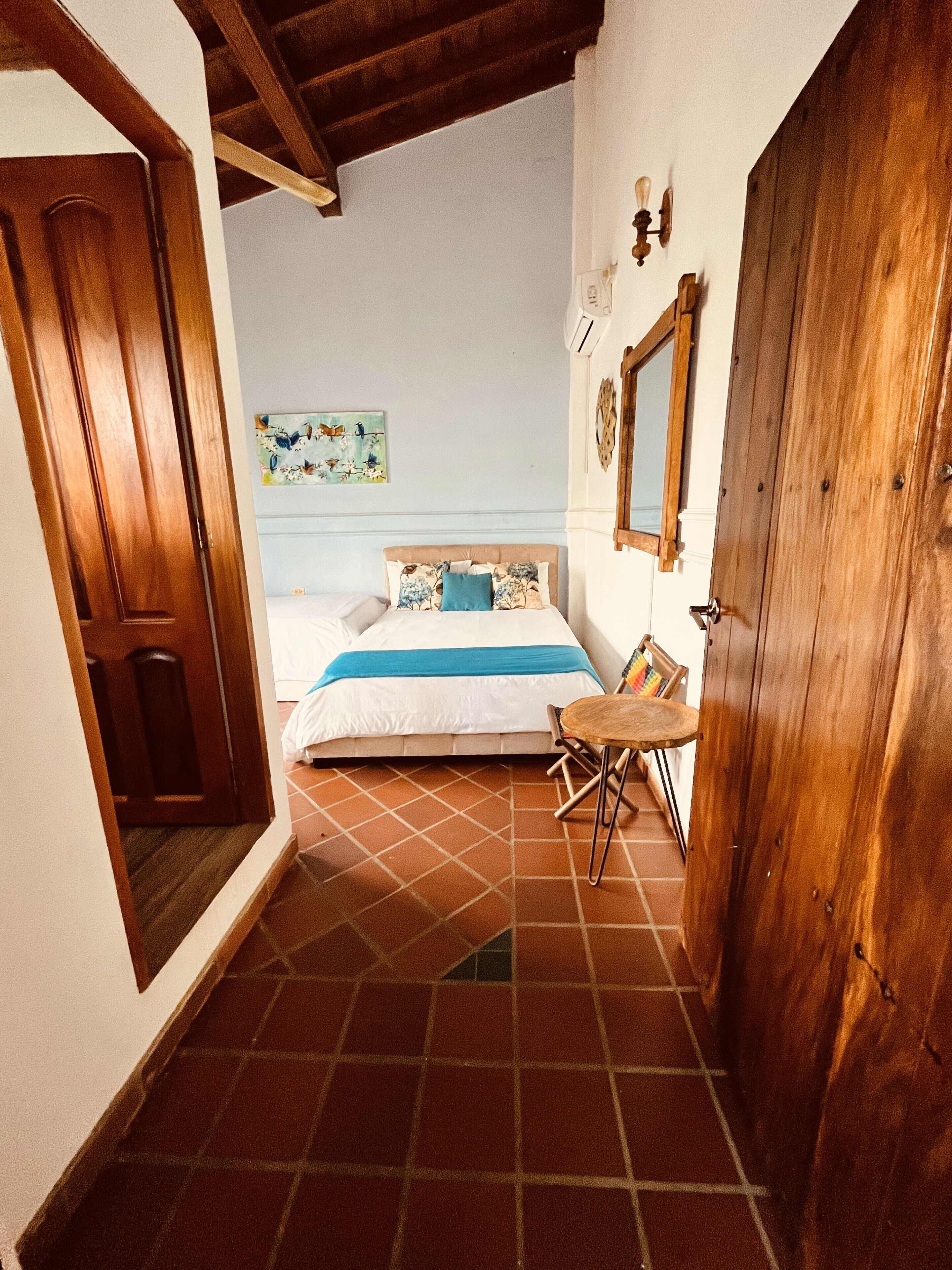 A bedroom with a bed, a small table, and a chair. There is a door on the left and a wooden door on the right.