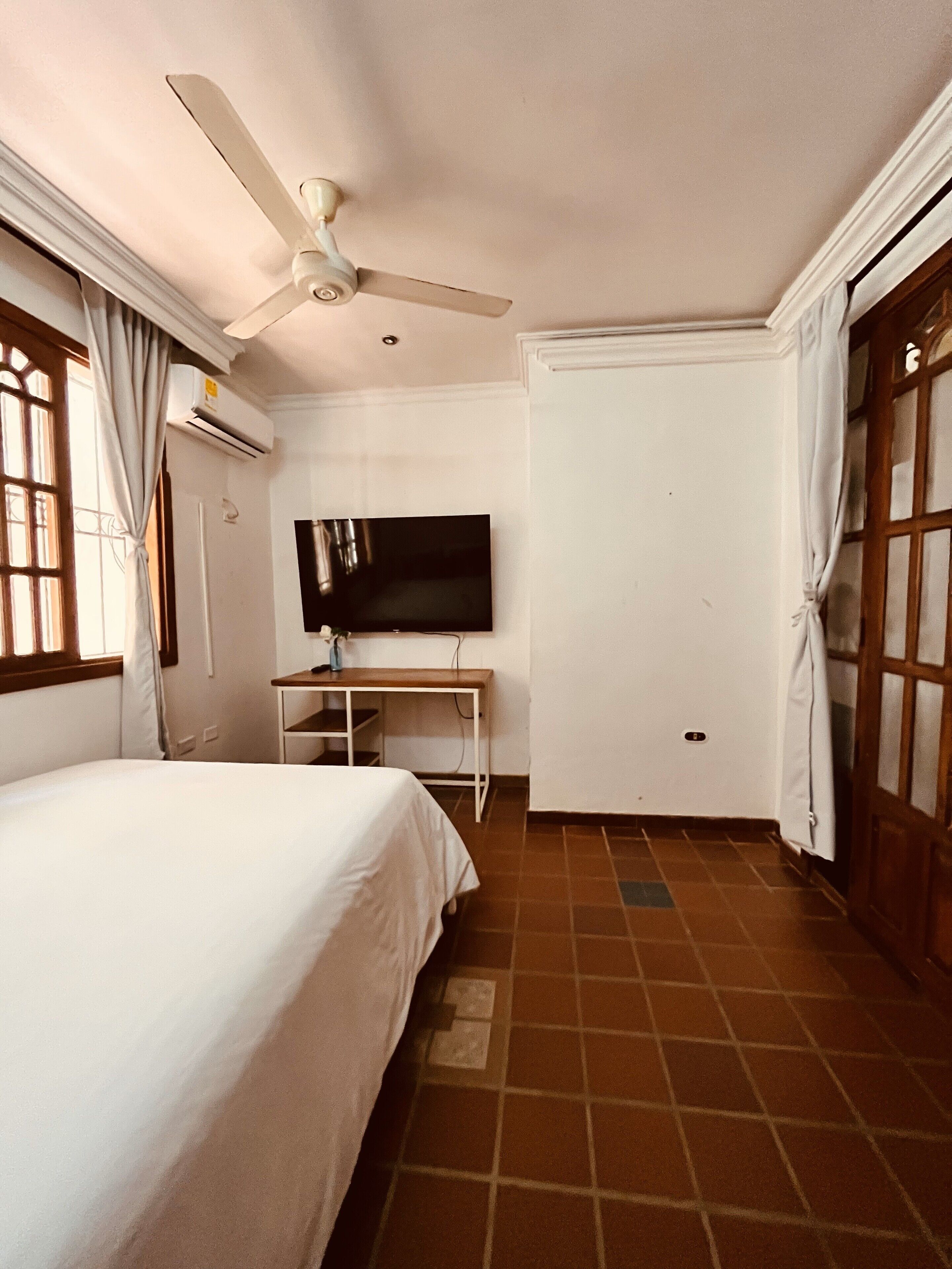 A hotel room with a bed, a desk with a TV, a ceiling fan, and a door with a window.