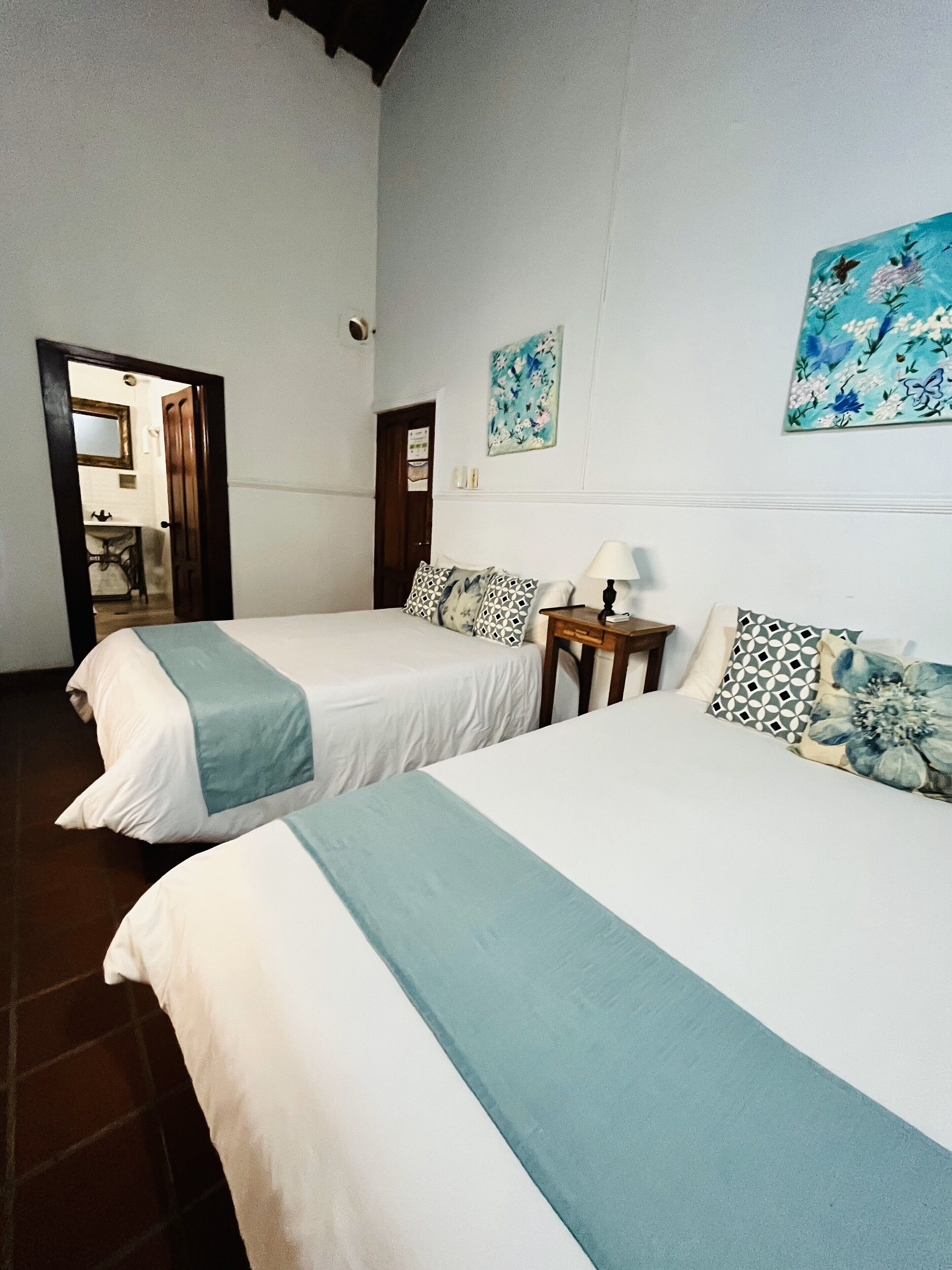 Dos camas individuales con cabeceras y pies de cama a juego, una mesita de noche con lámpara, y una obra de arte floral en la pared.